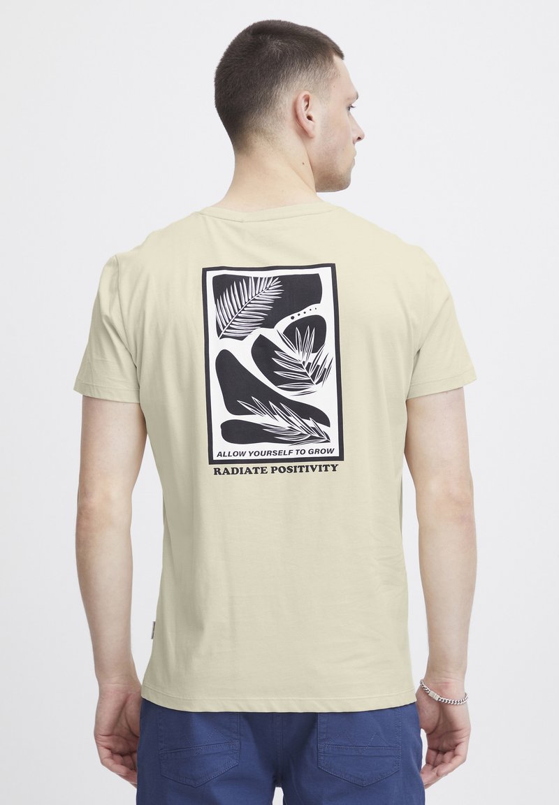 Lys beige t-skjorte med en svart grafisk design på baksiden med blader og teksten "TILLAT DEG SELV Å VOKSE" og "UTSTRÅLE POSITIVITET."