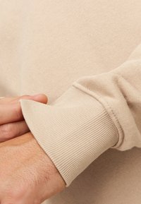 Polsino di felpa beige con texture a coste e finitura liscia. Il tessuto appare morbido e leggermente elastico, delineando una vestibilità rilassata.