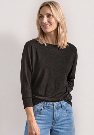 Lyshåret kvinde smilende, iført en sort langærmet skjorte og lyseblå jeans, står mod en ensfarvet lys baggrund.