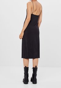 Robe noire midi avec des bretelles fines, silhouette ajustée et texture lisse. Associée à des bottines noires à talons épais et à fermetures éclair.