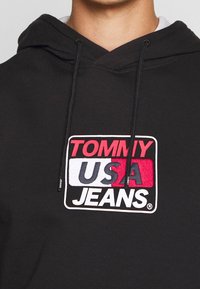 Zwarte hoodie met een kangoeroezak, met een opvallend geborduurd logo in rood, wit en blauw met de tekst "TOMMY USA JEANS." Met trekkoord in de capuchon.