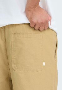 Beige Hose aus strukturiertem Stoff, mit einer großen Hintertasche und einem kleinen Logoanhänger an der Tasche. Der Bund sitzt über der Tasche.