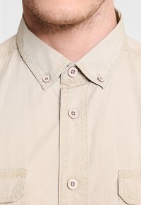 Camicia beige con bottoni a pressione e colletto classico, dotata di due tasche frontali e bottoni rosa. Realizzata in tessuto leggero con una texture liscia.