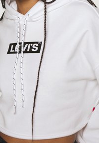Sudadera recortada blanca con el logo negro de "LEVI'S". Presenta cordones con rayas negras y un dobladillo acanalado. Textura suave de algodón.
