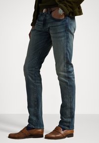 Vaqueros ajustados de mezclilla en azul medio, con un sutil efecto desgastado. Combinados con mocasines marrones y una chaqueta texturizada color oliva. Accesorios con un reloj.