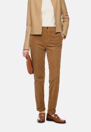 Person wearing tan jacket, brown corduroy pants, brown loafers, holding a brown clutch, standing against a plain background.