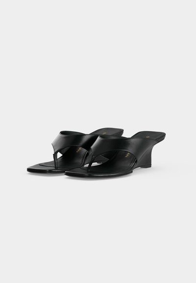 Sandales noires compensées en forme de tongs avec larges lanières en cuir et orteils découverts, présentées en angle sur un fond gris clair uni.