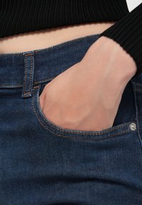 Main portant une chemise noire à manches longues rentrée dans un jean bleu en denim avec la main partiellement dans la poche avant.