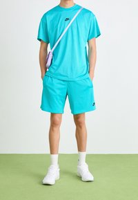 Osoba v tyrkysovém síťovaném tričku a šortkách Nike, s bílými teniskami a malou levandulovou přes rameno taškou, stojící na zelené podlaze.