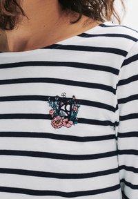 Marinblå och vitrandig långärmad skjorta med en blomsterbroderad design med en centralbokstav, mot en mjuk tygtextur.