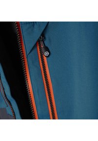 Blå stoffjakke med en kontrasterende oransje glidelås og svart trekk, med synlige sømmer og teksturert innvendig fôr.