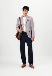 Hellgrauer Blazer mit einer Einstecktuch, weißem Hemd, marineblauen Hosen und braunen Lederschuhen. Braune Schultertasche und neutraler Hintergrund.