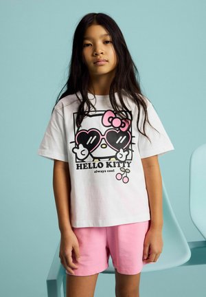 Young girl with long dark hair wears a white Hello Kitty t-shirt with heart-shaped sunglasses design and pink shorts, standing indoors.