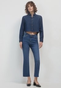 Camicia in denim blu scuro a maniche corte con chiusura frontale a bottoni, indossata con jeans blu a vita alta con gamba svasata e leggero strappo all'orlo.