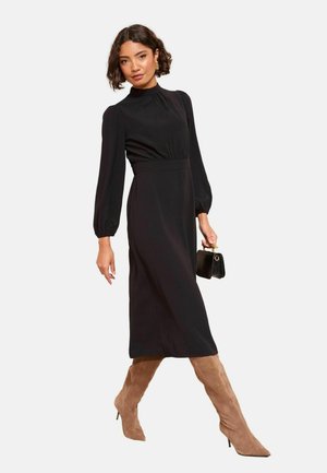 Schwarzes Midi-Kleid mit hohem Kragen, langen Puffärmeln und tailliertem Schnitt. Das Modell trägt taupefarbene Wildleder-Stiefel bis zum Knie und hält eine kleine schwarze Handtasche.