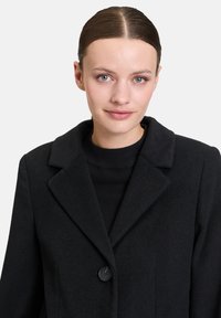 Schwarzer Wollmischungs-Mantel mit Reverskragen, zusätzlichem schwarzen Knopfverschluss und langen Ärmeln. Glatte Textur und zeitloses Design.