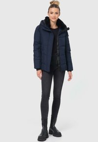 Marineblauwe pufferjack met een faux fur-voering in de capuchon, knoopsluiting aan de voorkant en ritszakken, gecombineerd met zwarte jeans en stevige zwarte laarzen.