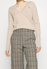 Pull beige en v avec manches longues, arborant un petit emblème doré sur la poitrine, associé à un pantalon large écossais brun.
