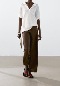 Weißes, gestricktes Poloshirt mit hochgekrempelten Ärmeln, kombiniert mit braunen Hosen mit weitem Bein. Schwarze und rote Flip-Flops. Clutch in der Hand; glatter, strukturierter Boden.