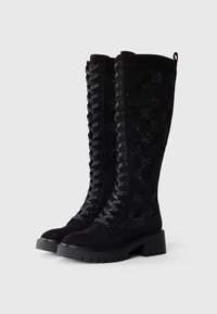 Black suede knee-high boots featuring a lace-up front, patterned panels, and a chunky black heel. Textured surface with cut-out details.