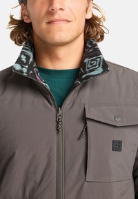 Chaqueta gris con cremallera y cuello de forro polar estampado, bolsillo de presión y costuras horizontales sutiles en el cuerpo. Presenta un logo en la parte inferior delantera.