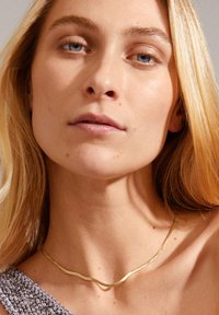 Gold wavy necklace resting on skin, featuring a smooth texture and sleek design. The model has long blonde hair and light skin tone.