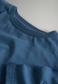 Kék, kerek nyakú póló kis mellzsebbel, egy világosszürke felületen laposan elhelyezve.