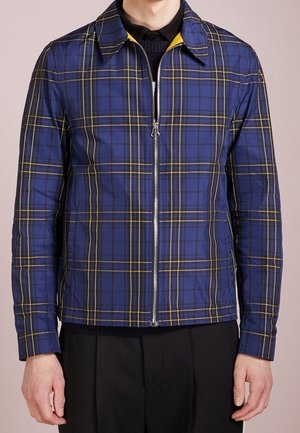 Niebieska kurtka w kratę na zamek błyskawiczny z żółtymi i czarnymi liniami, noszona na czarnej koszuli z kołnierzykiem i czarnych spodniach, pokazana od ramion do bioder.