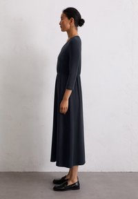Czarna, długosleeve, sukienka w kształcie litery A z dopasowanym wcięciem w talii i okrągłym dekoltem. Modelka nosi czarne mokasyny, stojąc na neutralnym tle.