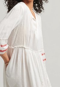 Biała sukienka z teksturowanej tkaniny z kropkowanym wzorem. Posiada czerwone haftowane akcenty na rękawach oraz ściągacz w talii z frędzlami.