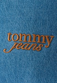Modrá rifľovina s hladkým povrchom, na ktorej je vyšitý text "tommy jeans" svetlohnedým kurzívnym písmom.
