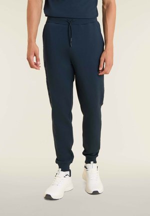 Jogges navy en tissu doux avec une taille élastique, des rayures latérales et des poignets côtelés, associés à des chaussures de sport blanches.