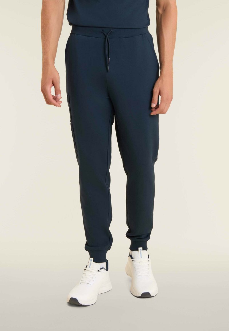 Pantalones de chándal azul marino fabricados en un tejido suave, con una cinturilla elástica, rayas laterales y puños de canalé, combinados con zapatillas deportivas blancas.