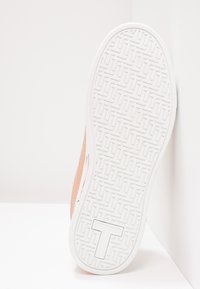 Zapatilla con un material superior suave en color durazno, suela de goma blanca texturizada y un diseño de agarre con patrón. El lateral presenta un sutil acento rosa.