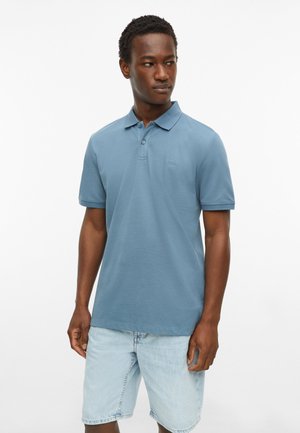 Man die een blauw poloshirt met korte mouwen en lichte spijkershorts draagt, kijkt naar rechts tegen een effen witte achtergrond.