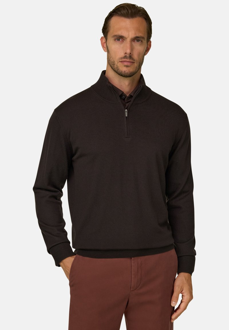Homme aux cheveux courts et barbe porte un pull noir à fermeture éclair quart sur une chemise à col et un pantalon marron, debout avec une main dans la poche.