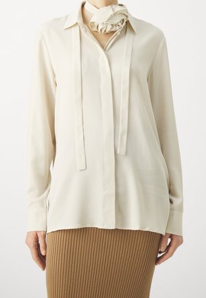 Femme portant un chemisier crème à manches longues avec un foulard noué au cou et une jupe côtelée de couleur camel.