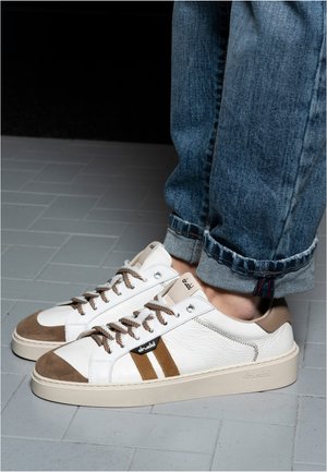 Person wearing white and brown low-top sneakers with striped laces and rolled-up blue jeans standing on tiled floor.