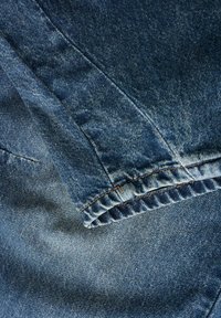 Denim tkanina v srednje modri barvi, z gladko teksturo in kontrastnim šivanjem. Posnetek od blizu zložene in plastične dele.