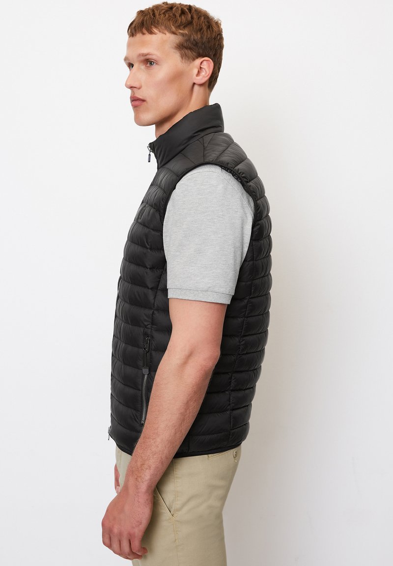 Gilet matelassé noir avec un col haut, des poches zippées et un design léger, porté par-dessus une chemise à manches courtes gris clair.