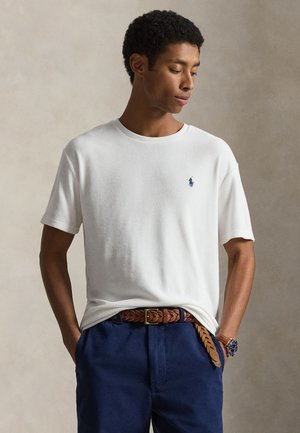 Man draagt een wit poloshirt met korte mouwen, een gevlochten bruine leren riem en donkerblauwe broek, kijkt naar beneden met handen in de zakken.