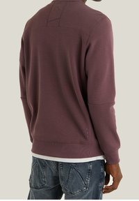 Felpa viola con scollo a girocollo, tessuto strutturato e dettagli di cuciture visibili. Indossata sopra una maglietta bianca e abbinata a jeans scuri.