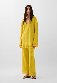 Geel gebreid tweedelig outfit met een lange mouwen top met franjes en een broek met wijde pijpen, versierd met textuurpatronen en gefranjerde zomen.