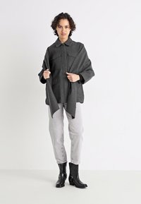 Giacca grigia in tessuto intrecciato con due tasche sul petto, indossata sopra una maglietta nera a maniche lunghe. Abbinata a jeans grigio chiaro e ankle boot neri.