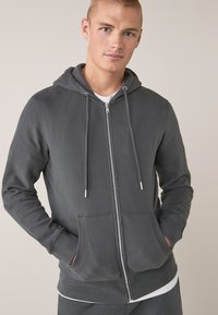 Sudadera gris con cremallera, hecha de un tejido suave, que cuenta con una capucha con cordones, bolsillos frontales y detalles en hardware plateado. Diseño simple con una textura suave.