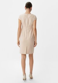 Beiges Kleid mit kurzen Ärmeln, taillierter Silhouette, Reißverschluss im Rücken und einem Schlitz im Rücken für Bewegungsfreiheit, kombiniert mit beigen Absätzen.
