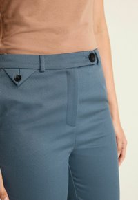 Närbild av en person som bär blå hög midja byxor med svarta knappar och en inskjuten beige ärmlös topp mot en enkel bakgrund.