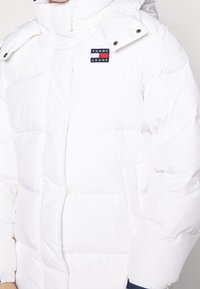 Vit puffjacka med hög krage, avtagbar huva och Tommy Jeans-logotyp. Tyget är slätt med en quiltad textur.