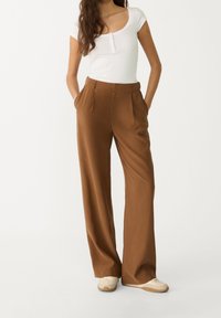 Pantalons larges marron avec une texture lisse, dotés de plis avant et de poches, associés à un haut blanc à encolure dégagée et à des baskets beiges.