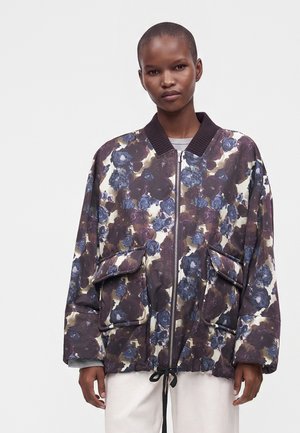 Personne portant une veste ample à motif floral avec de grandes poches et une fermeture éclair, associée à un pantalon clair sur un fond uni.